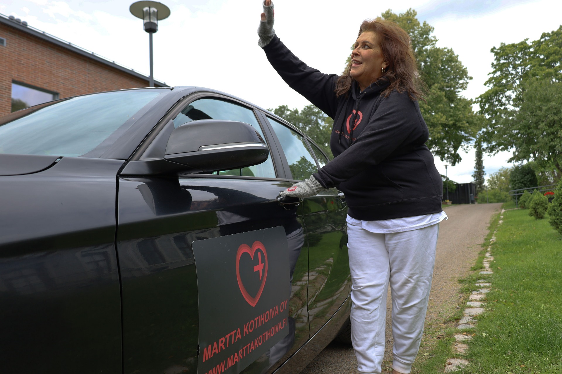 Kotihoitaja nostaa kättään. Menossa autoon, ulkona pihatiellä, taustalla puita ja tiilirakennus.